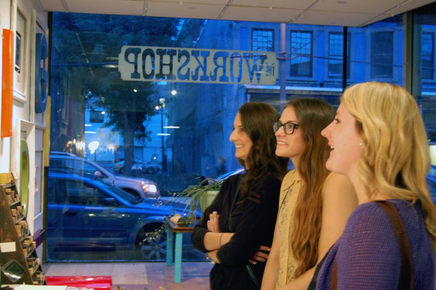 Meredith Harding and Ashley Kujat, senior graphic design majors, and Emily Mouillesseaux look at art from the CMYK showcase at The Workshop.